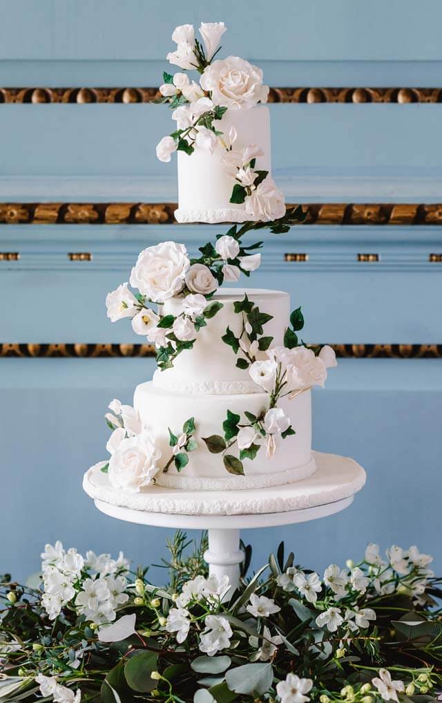 3 tier white wedding cake with floating tier between top and middle tiers. Sugar roses, lisianthus and dark ruscus cascading down from the top tier to the base